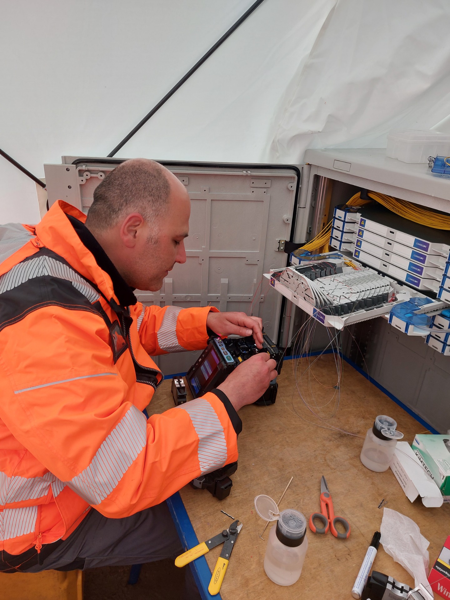 Technician in orange jacket works inside open equipment rack, using handheld device amid cables and components.